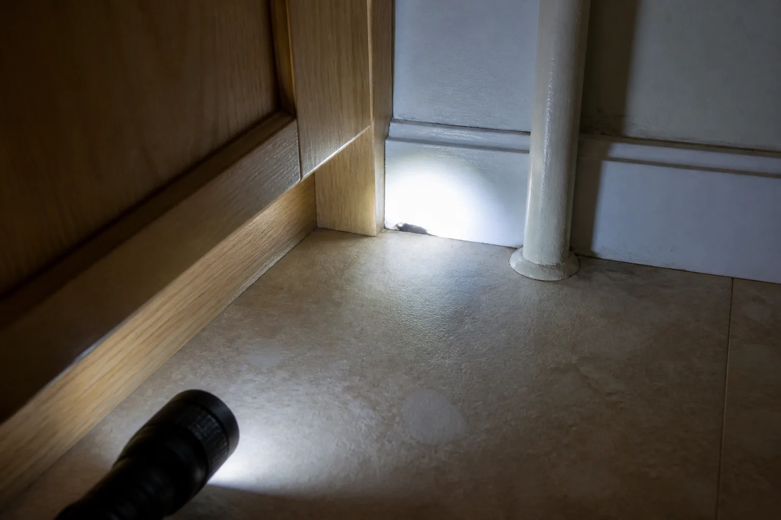 A flashlight aimed at a pencil-width gap near a cabinet and utility pipe