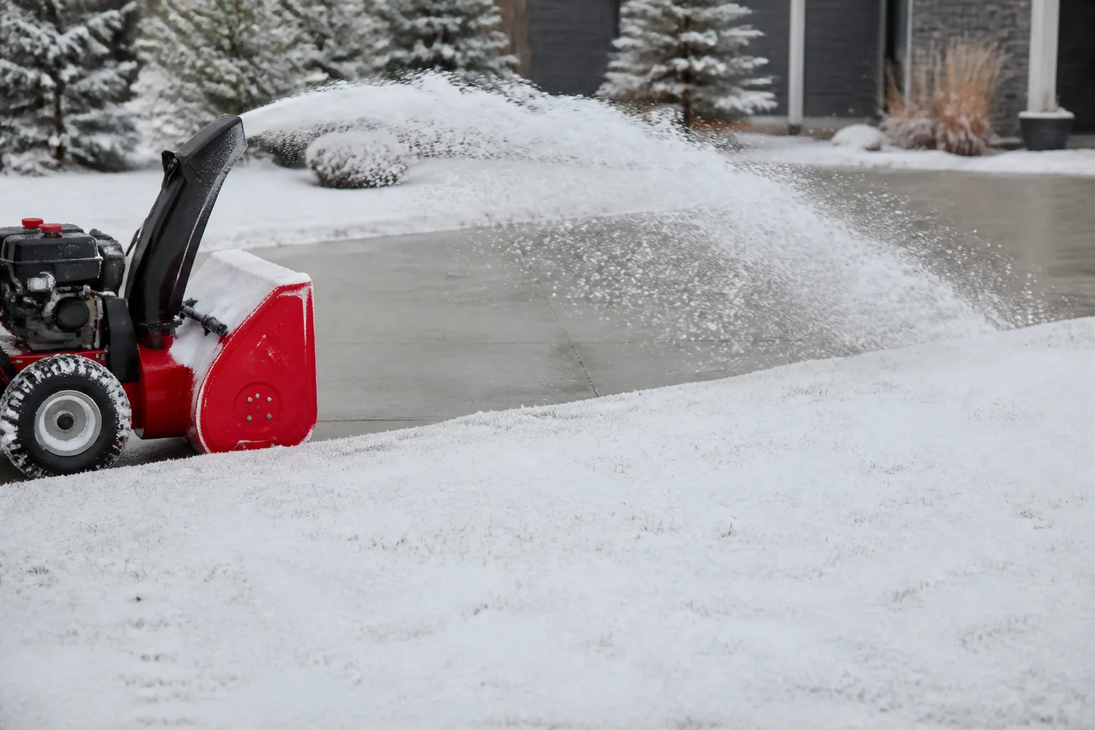 Snow blower clearing a driveway with discharge chute aimed away from the dormant lawn toward a paved apron